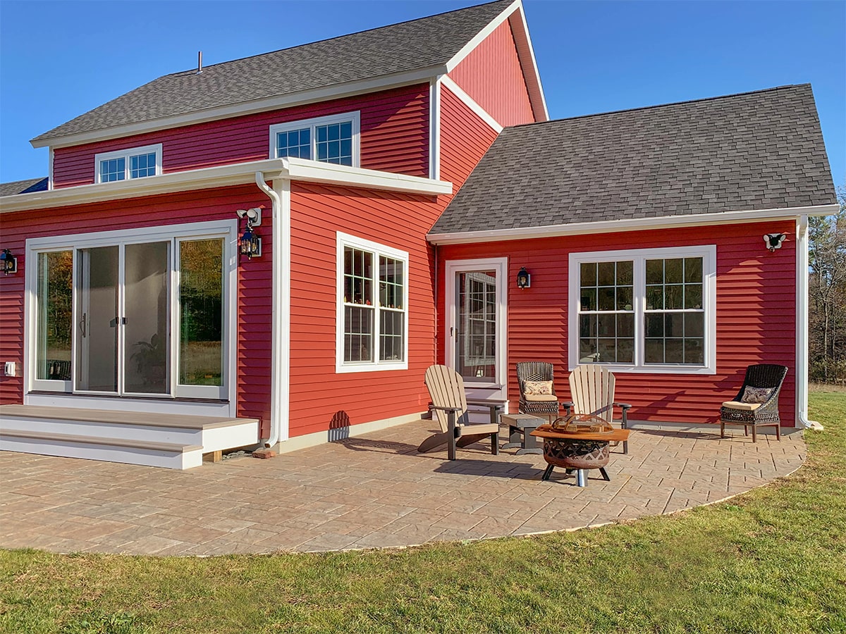 Exterior view of a red modern farmhouse with white trim, large windows and sliding doors opening onto a paved patio with outdoor chairs and clear blue sky