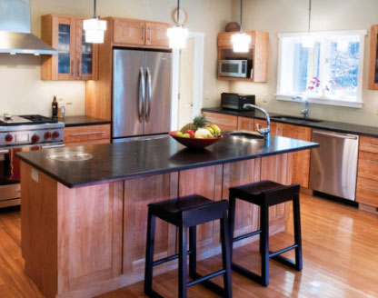 Modern kitchen with large central island featuring dark granite countertop, wooden cabinets, three pendant lights, two bar stools, stainless steel appliances, and fruit bowl
