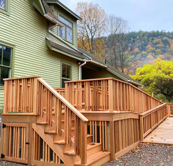 Wooden exterior deck with stairs and long ramp featuring vertical slat railings, attached to green-sided house, overlooking autumn forested hillside