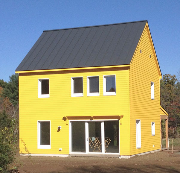 Sunny yellow exterior home with black metal roofing, symmetrical upper windows, expansive lower glass doors opening to patio area, and natural wooded setting